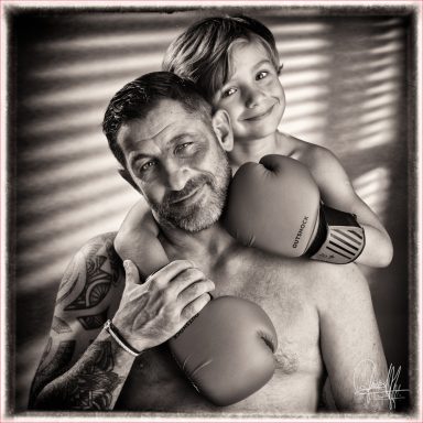 Jolis portrait de famille en noir et blanc d'un petit garçon souriant avec des gants de boxe sur les épaules de son Papa.  