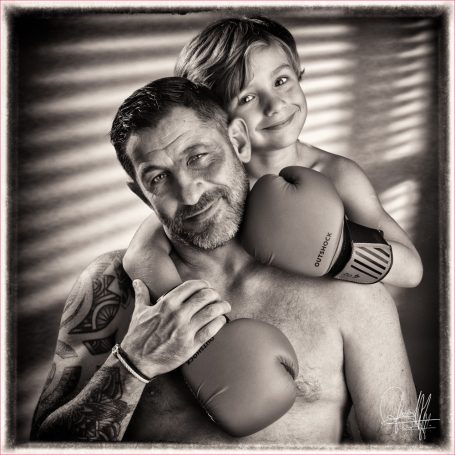 Portrait de famille en noir et blanc, petit garçon avec gants de boxe sur les épaules de son papa, moment complice